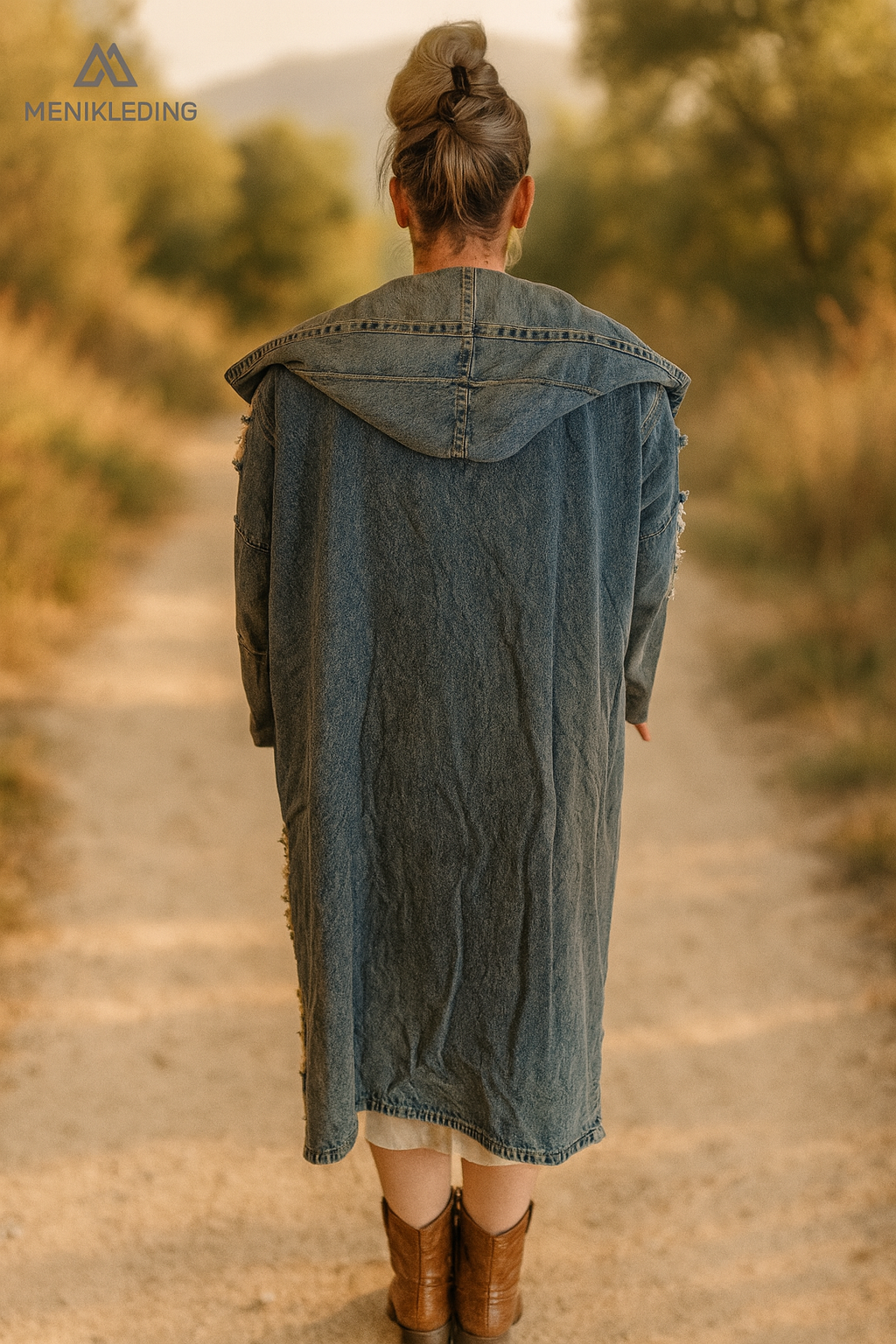 Achteraanzicht van een lange bohemian spijkerjas met capuchon van Menikleding, gemaakt van denim met een ruwe afwerking, gedragen door een vrouw met opgestoken haar en laarzen.